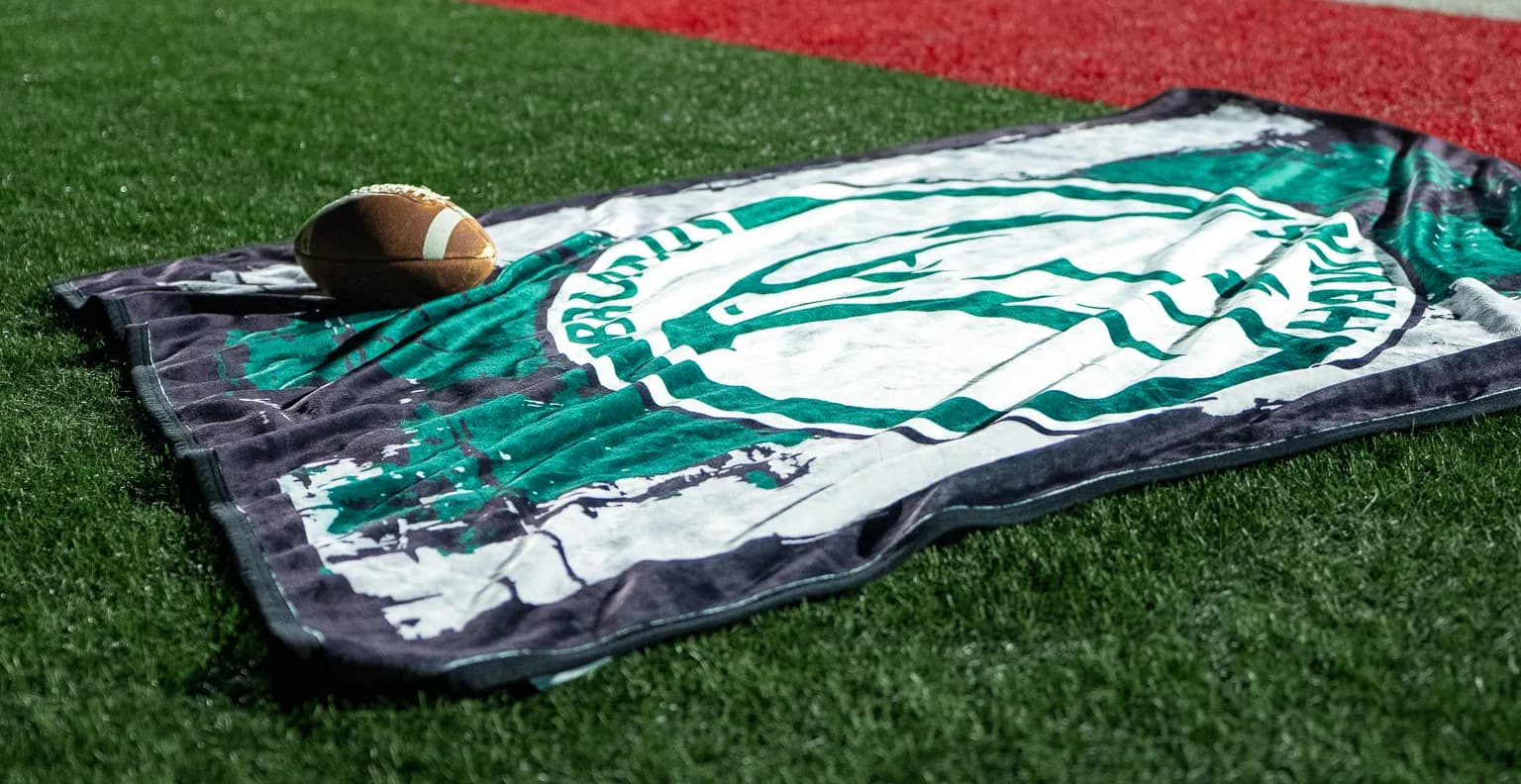 A green Bruins mascot blanket laid out in the endzone with a football, under the stadium lights at night