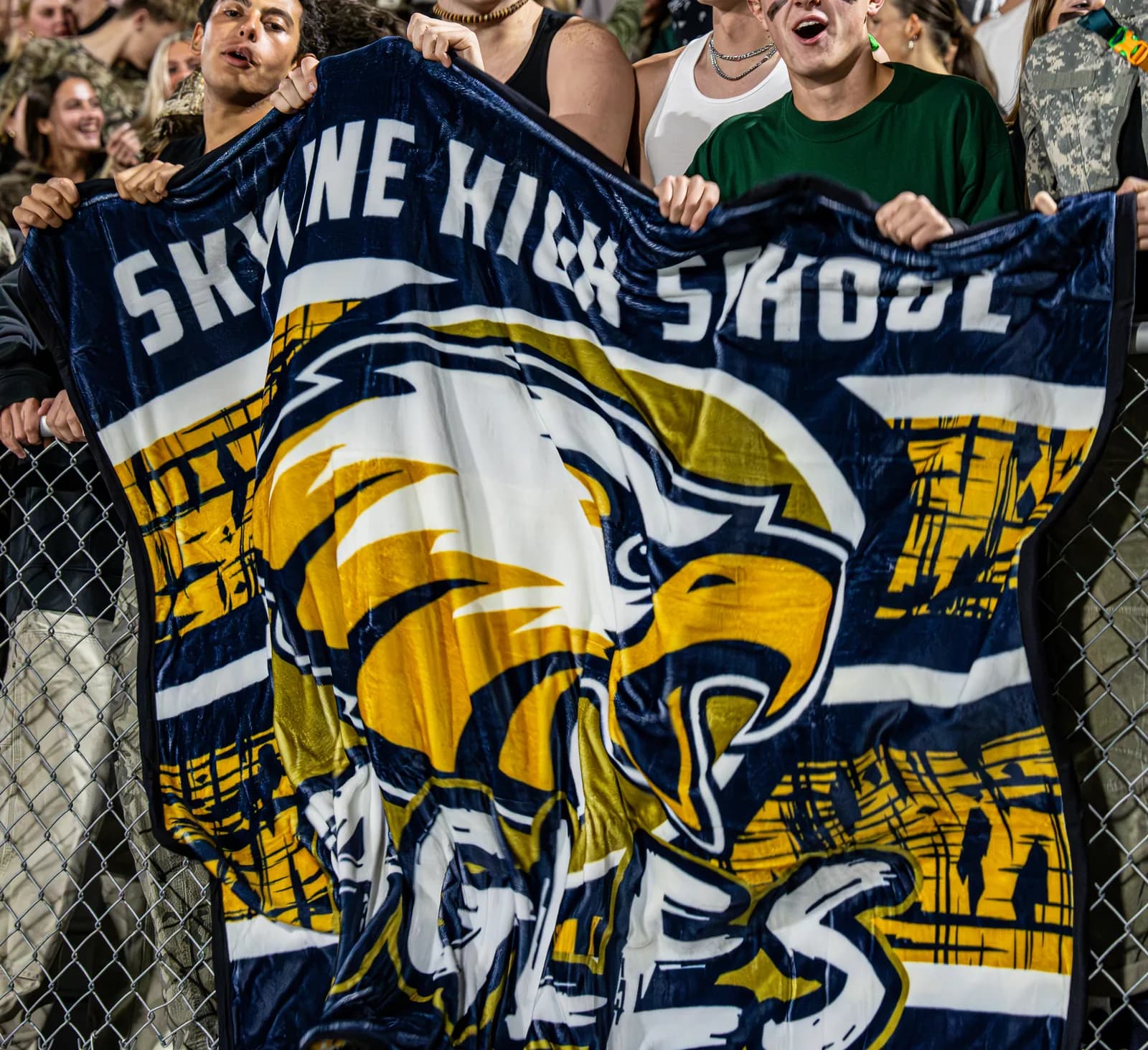 Students in the stands holding up their custom Skyline High School eagle mascot blanket at a night game